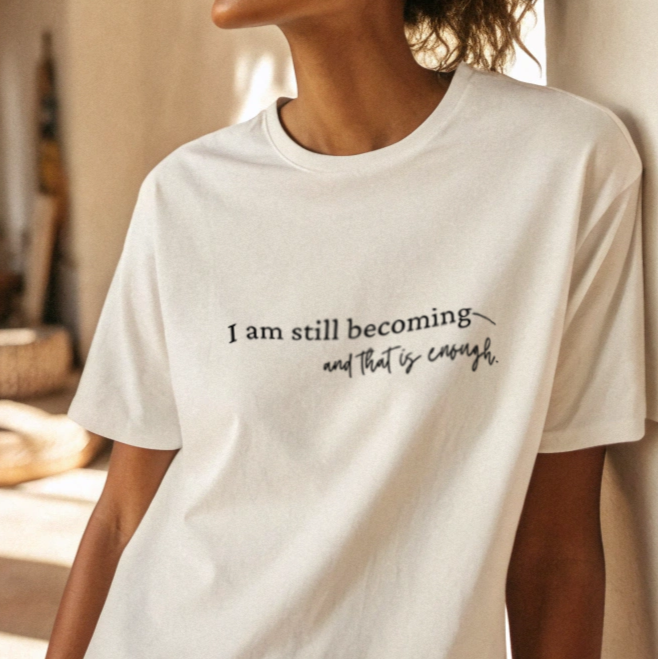 Person wearing a white t-shirt 'I am still becoming and that is enough' in a casual setting.